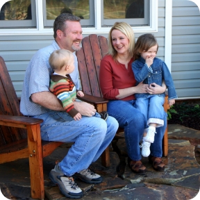 Couple sit outside on patio furniture. Each hold a child on their lap. 